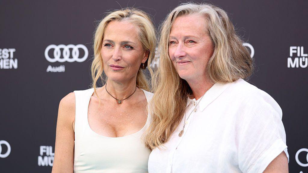 Actress Gillian Anderson on the left, wearing a sleeveless white dress with her blonde hair in a ponytail, looks away from the camera. She is stood next to author Raynor Winn, who has long and wavy fair hair and wears a short sleeved white shirt.