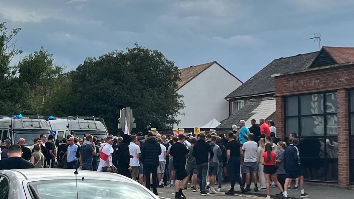 A large group of people walking and gathering outside buildings. Most are wearing black and one person has a St George's flag draped over their shoulders. There are two police vans visible behind the group in the background.
