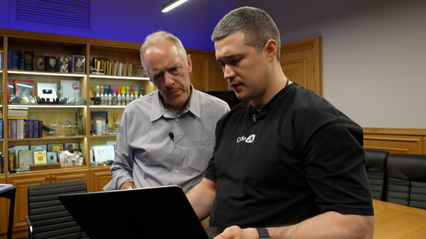 Two men stand looking at a laptop, one in the foreground in a black T-shirt, the other in a grey shirt
