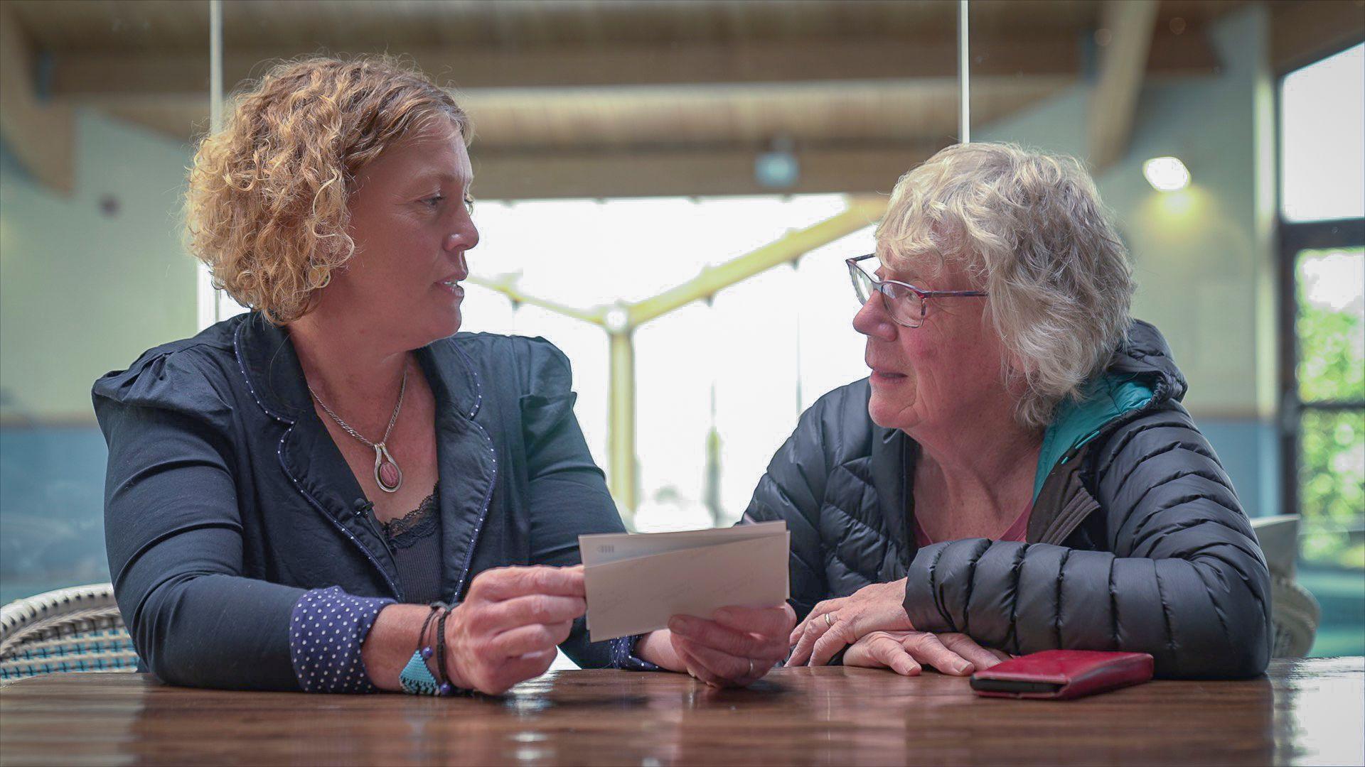 Two women sat at a table next to each other, looking at family photographs. They look at each other as they hold the photos.
The woman on the left is wearing a navy jacket and has short ginger curly hair. The women on the right has short grey hair, glasses and is wearing a dark coat.