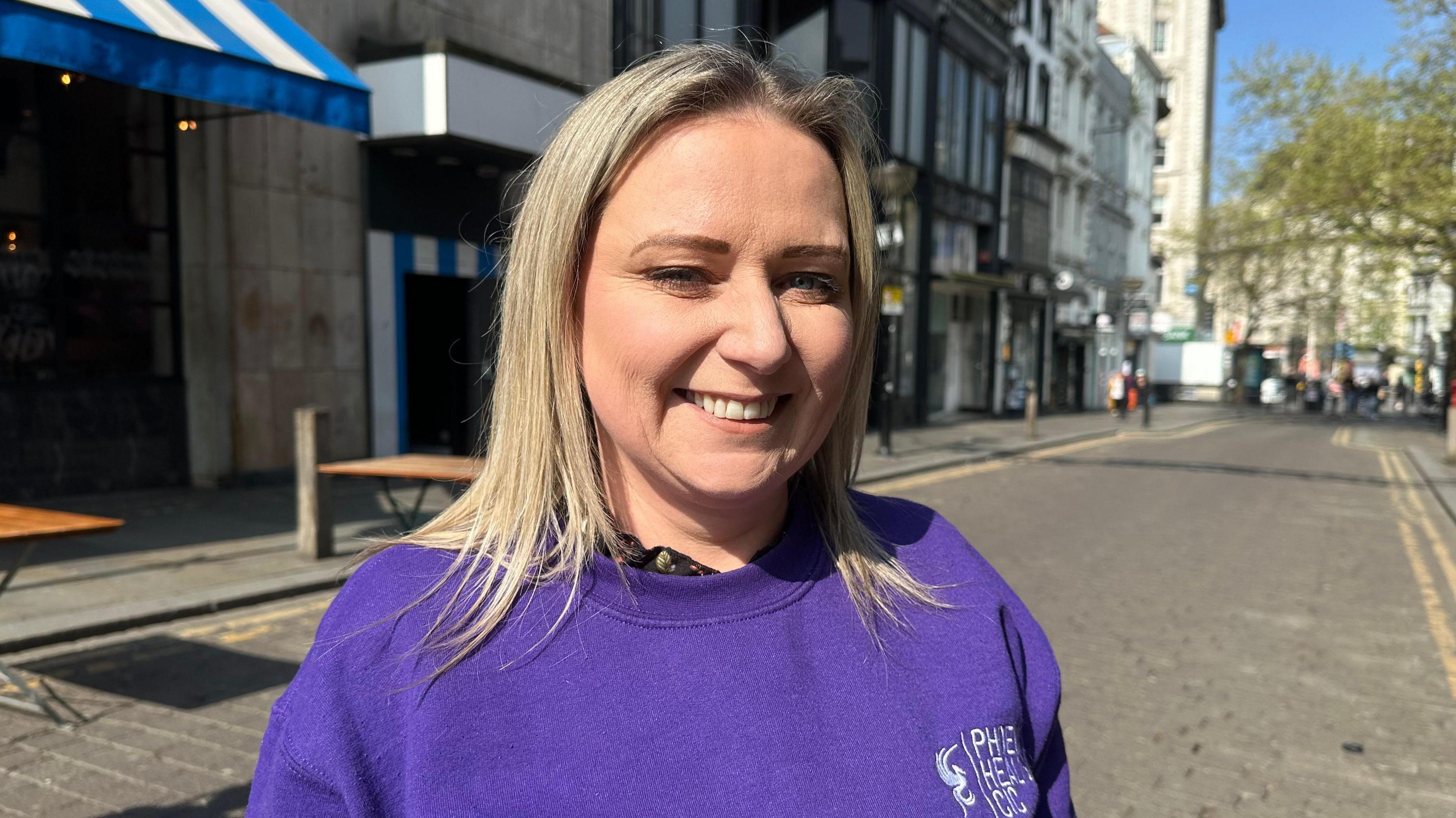 Jemma Stewart has blonde hair and is dressed in a purple sweatshirt with the logo of her organisation