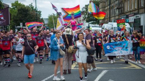 The Newcastle Pride march in 2024. Crowds of people are walking down the middle of the road towards the camera. They are wearing brightly coloured cloths and are waving rainbow flags. 