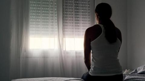 a stock image to represent domestic abuse. A woman with long hair ties back in a low ponytail, wearing a vest top is sat at the edge of a bed in a darkened room. She is facing the window, with a net curtain drawn. Sunlight is visible through the curtain.