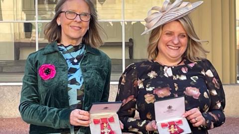 Carole Gould and Julie Devey hold up their OBE medals to the camera. They are both smiling and Carole is wearing a smart hat