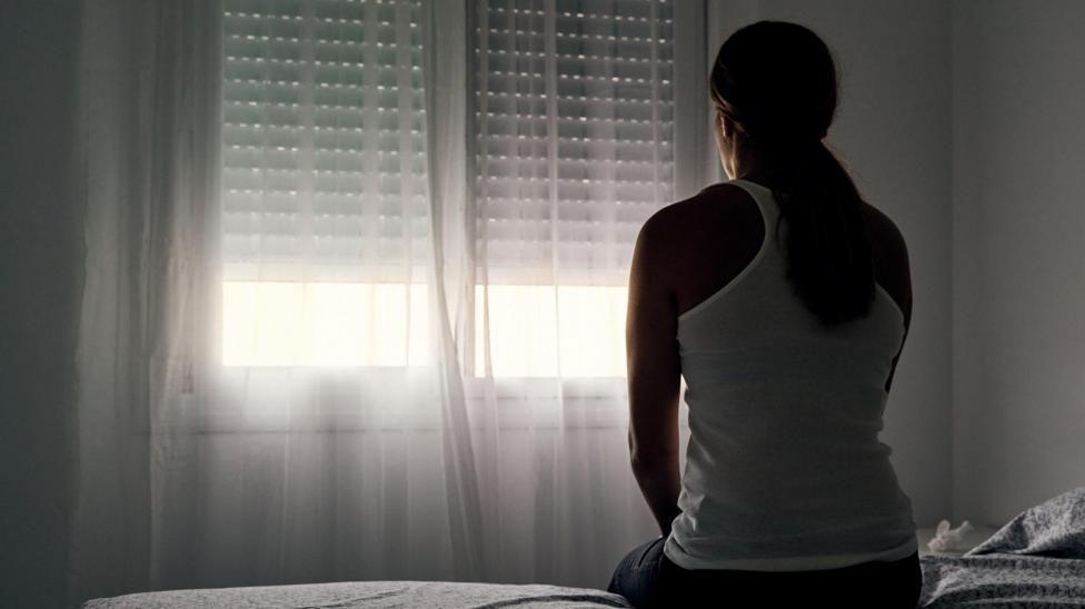 a stock image to represent domestic abuse. A woman with long hair ties back in a low ponytail, wearing a vest top is sat at the edge of a bed in a darkened room. She is facing the window, with a net curtain drawn. Sunlight is visible through the curtain.