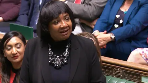 Diane Abbott stands in the House of Commons wearing a black outfit and a large statement necklace. She is surrounded by seated MPs dressed in various colors, including blue, grey, and red. The chamber’s green benches and ornate wooden paneling are visible in the background.