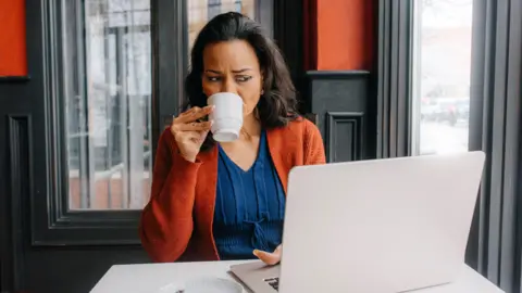 A woman is sipping from a white mug while working on her laptop at a cafe. She appears to be focused, surrounded by a warm interior with red and black accents.