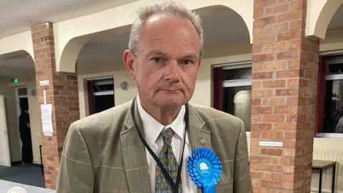 A man with short grey hair, a grey jacket, green tie and a blue Conservative Party rosette, stands in a hall with brick pillars and a white wall with windows behind him.